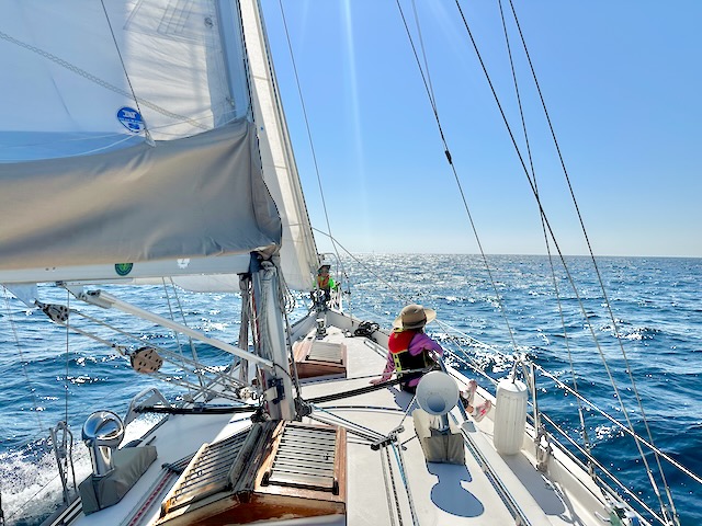 The view from Festina Lente's dodger.All three sails are deployed, and ahead is nothing but open water. One child is hanging out on the windward (starboard) side of the boat, while another hangs out up at the pulpit.